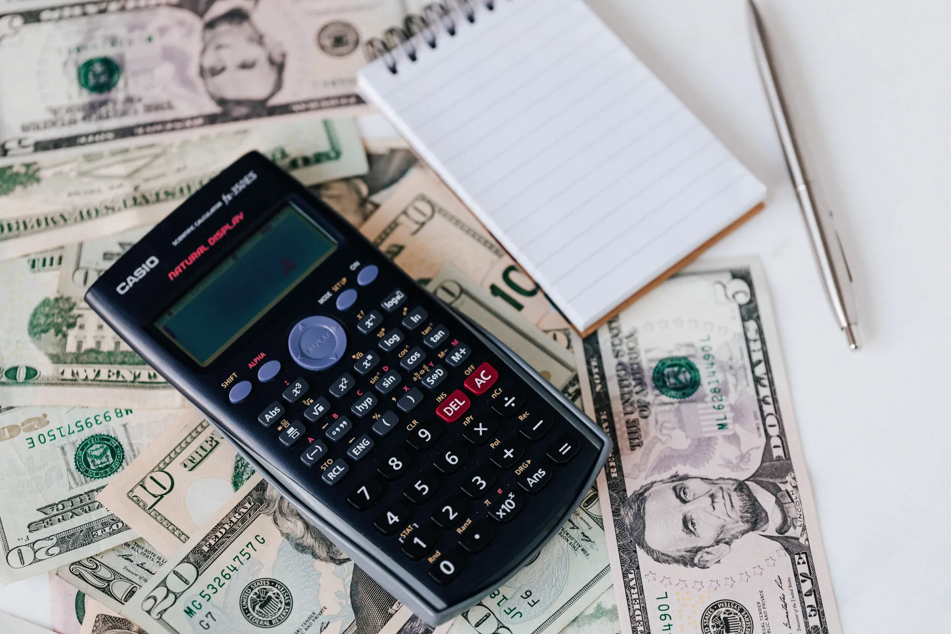 Calculator, blank notebook and pen arranged over US dollar bills.
