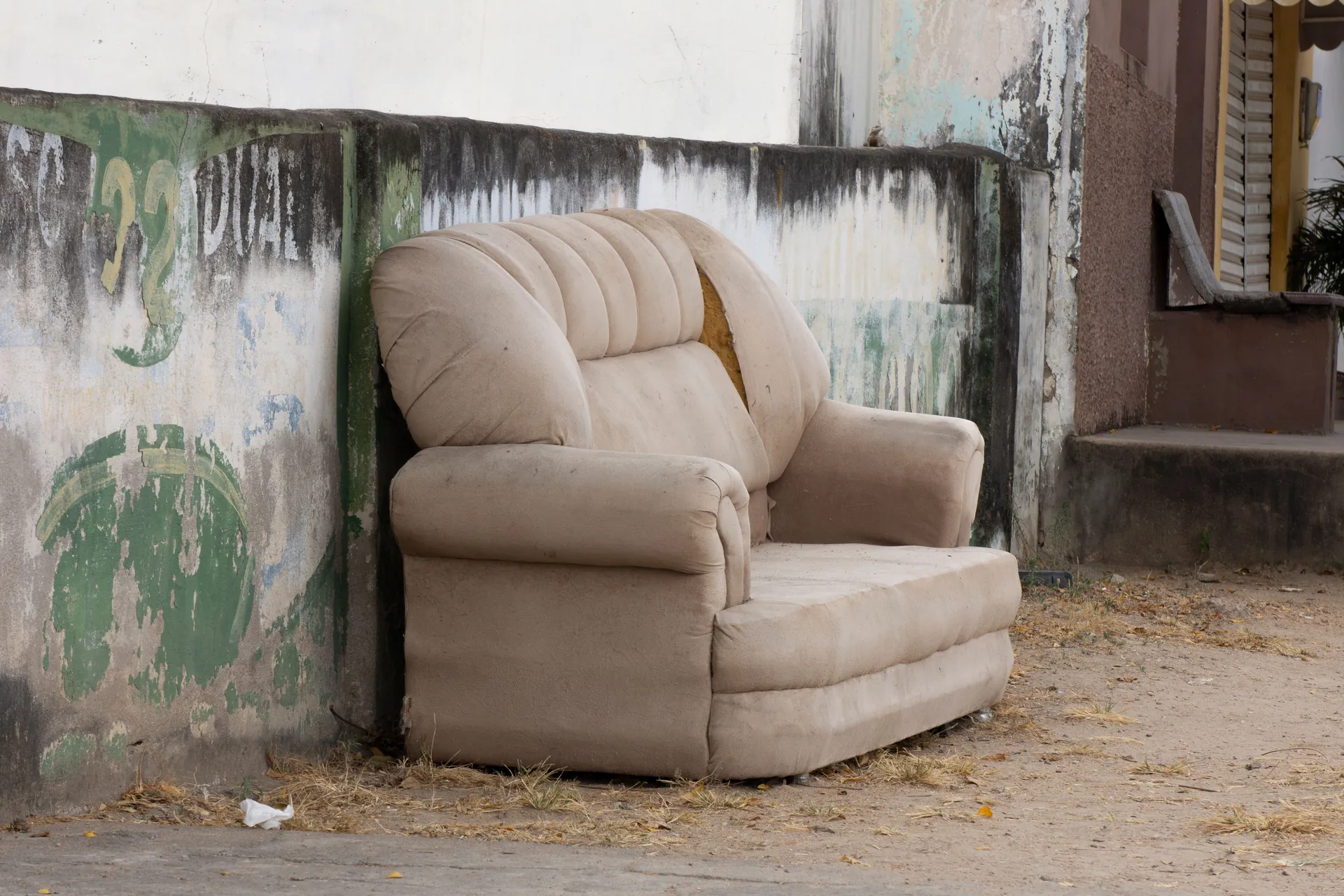 Discarded beige upholstered armchair beside a weathered outdoor wall.