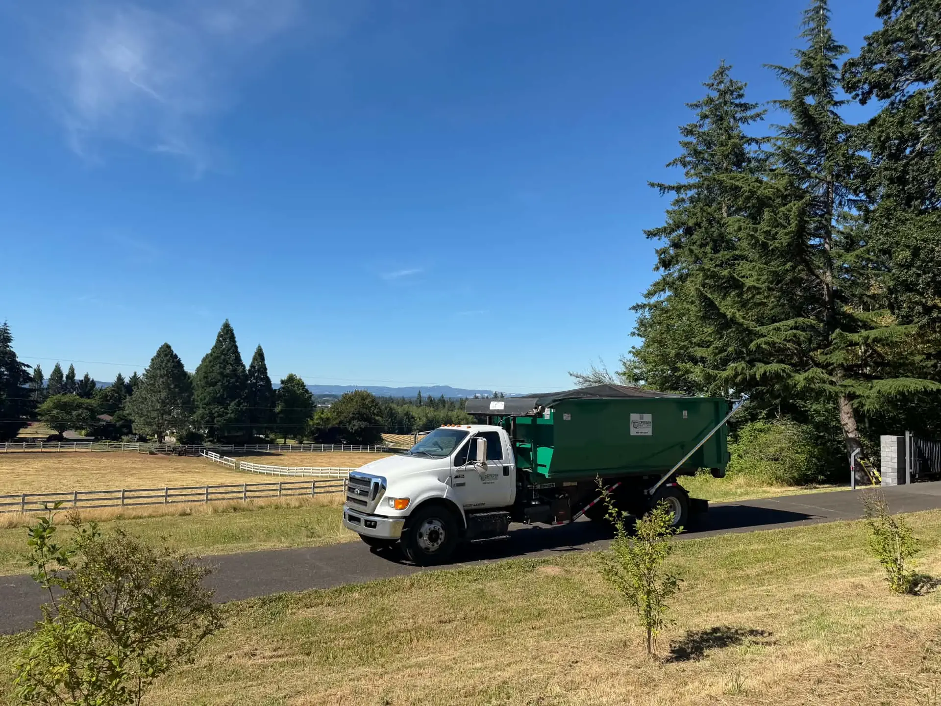 Oregon Recycling white truck carrying a green roll-off dumpster beside a pasture.