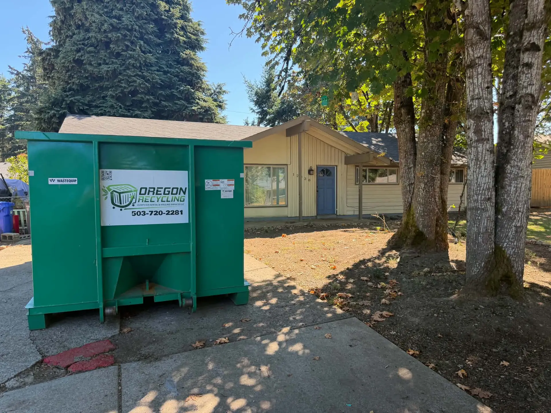 Green dumpster displaying the Oregon Recycling name and phone number beside a home.